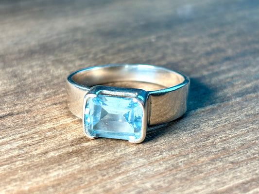 A silver ring featuring a rectangular light blue gemstone, resting on a textured wooden surface.