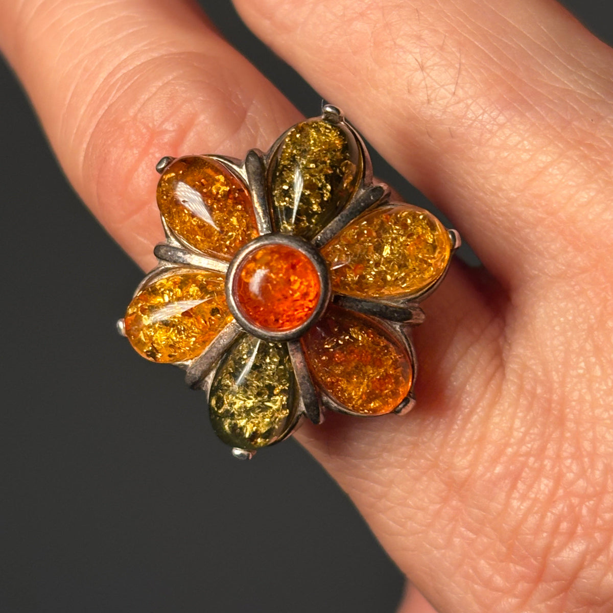 ring with six oval amber petals in honey, cognac, and green tones surrounding a round orange center, worn on a finger.