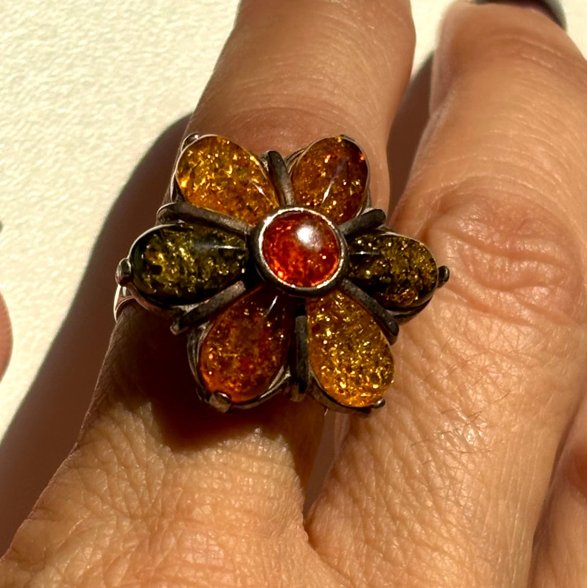 A close-up of a hand wearing a flower-shaped ring with amber and olive-green stones in a dark metal setting.