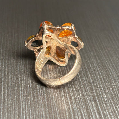 A silver ring featuring a floral cluster setting with amber-orange and yellow gemstones, photographed on a textured gray background.