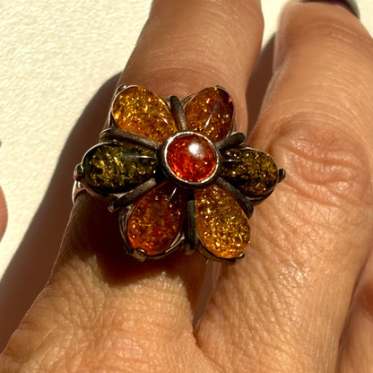 A close-up of a hand wearing a flower-shaped ring with amber and olive-green stones in a dark metal setting.