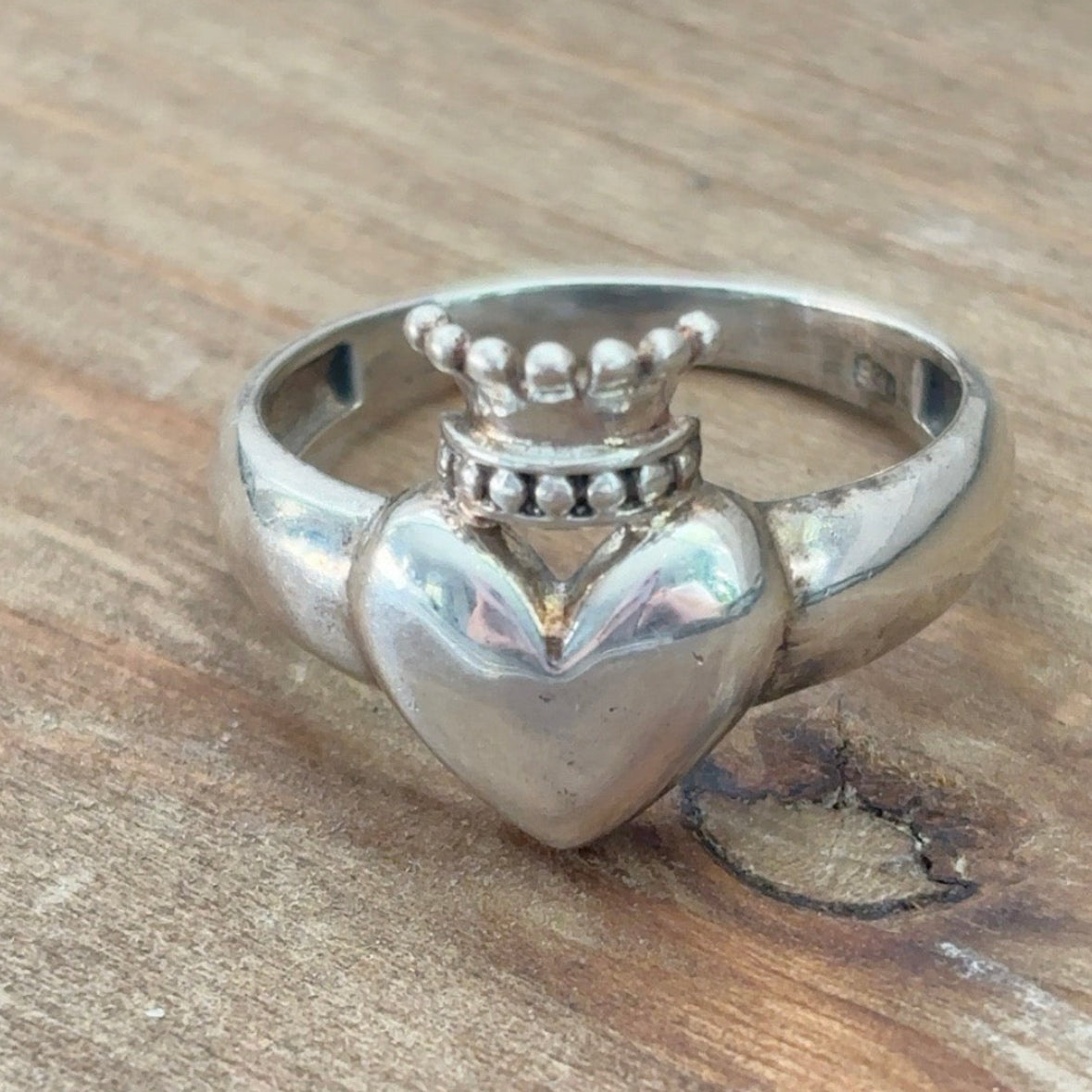 A silver heart-shaped ring with a crown on top, photographed on a rustic wooden surface.