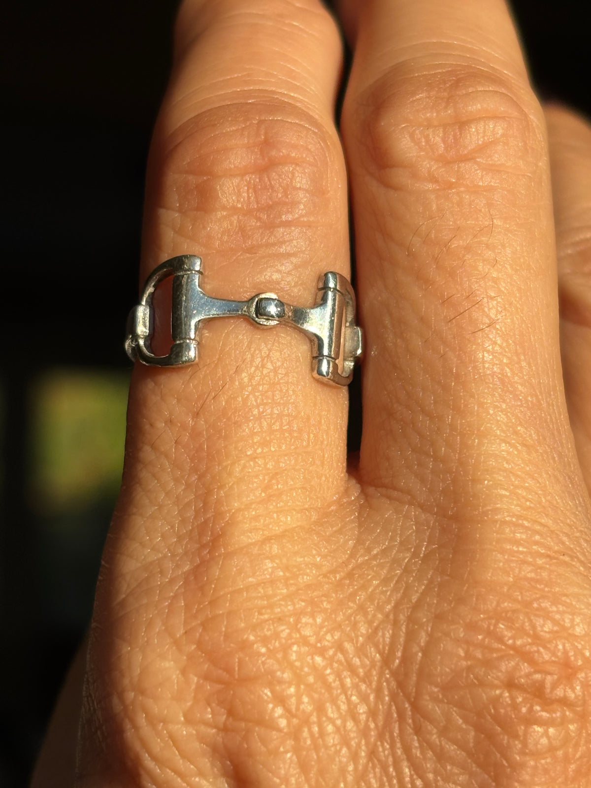 A sterling silver horsebit ring worn on a finger, shown in a close-up shot with warm, direct lighting against a dark background.