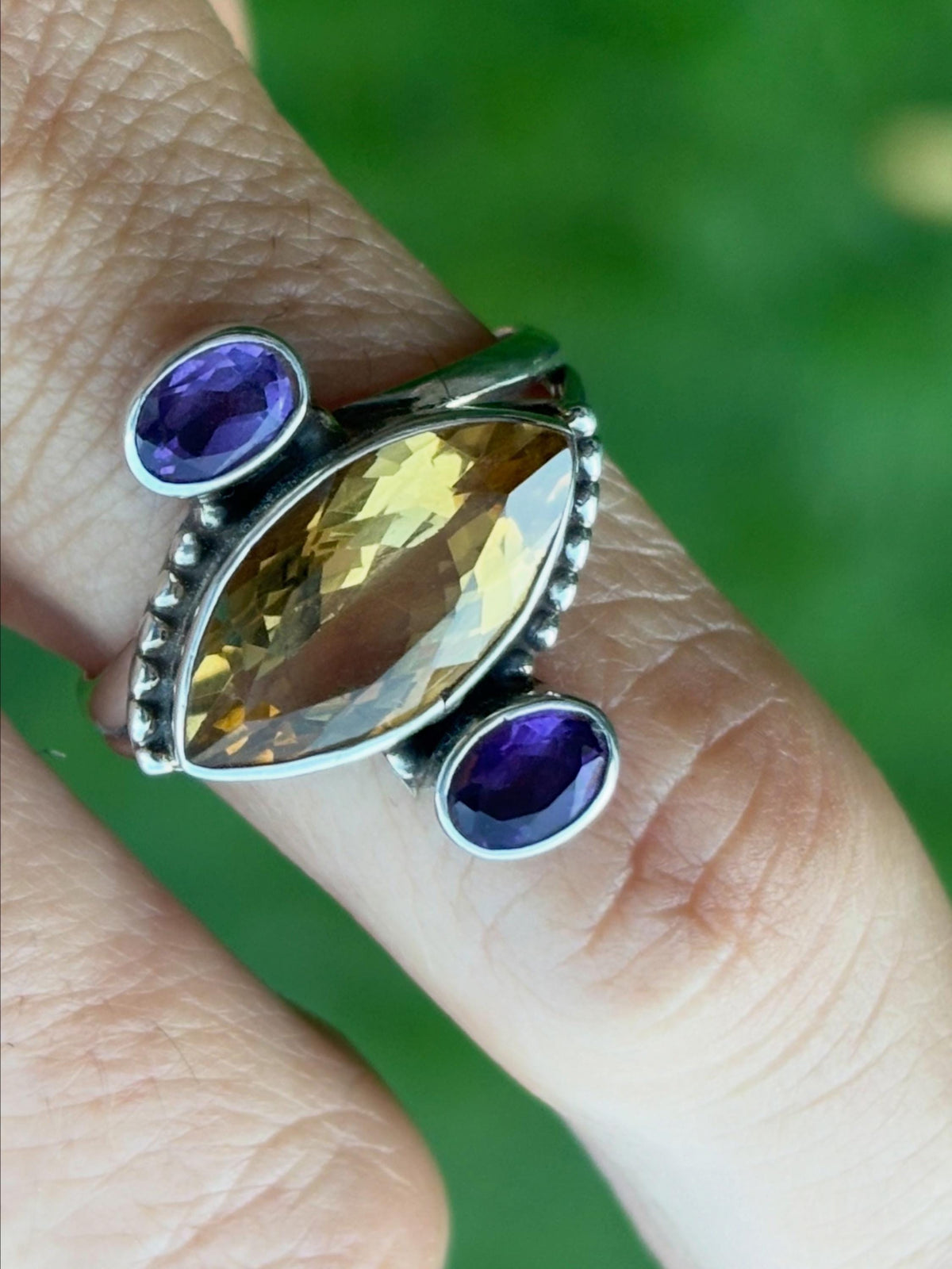 A close-up of a silver ring worn on a finger, featuring a large marquise-cut yellow gemstone in the center with two oval purple gemstones on either side.