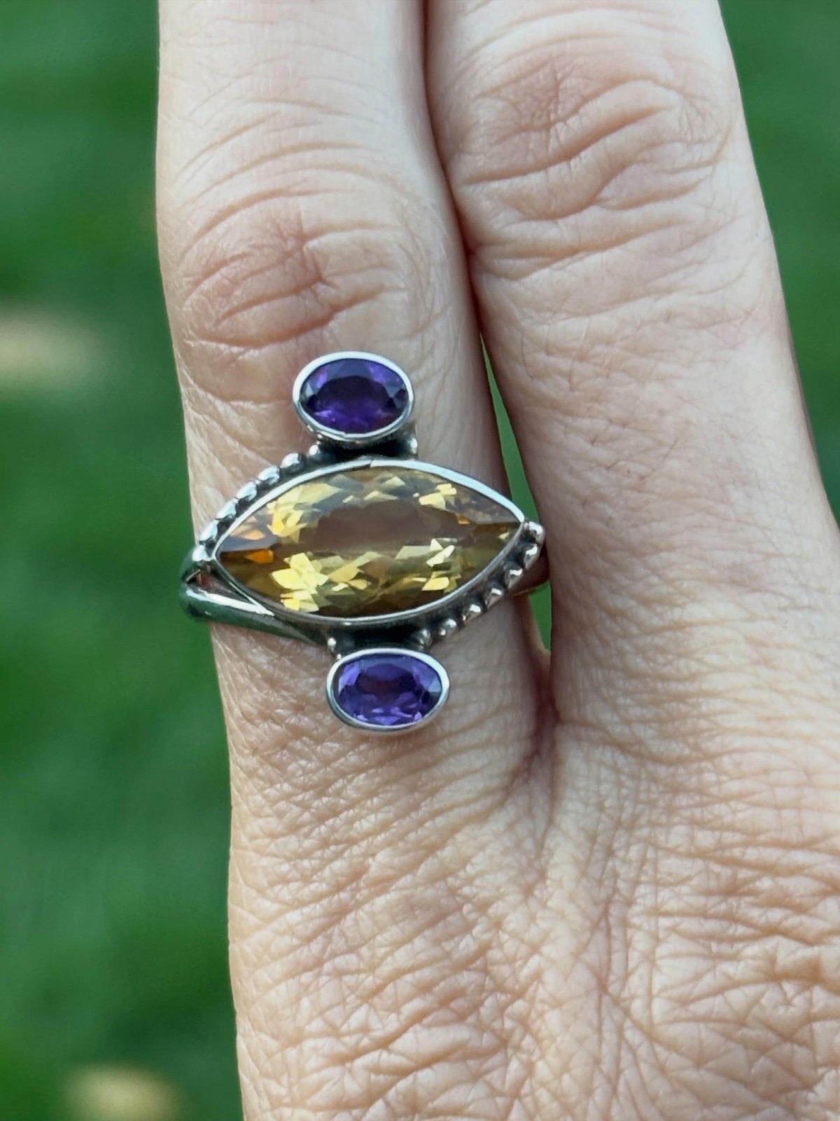 A close-up of a hand wearing a silver ring with a large marquise-cut yellow gemstone and two oval purple gemstones.